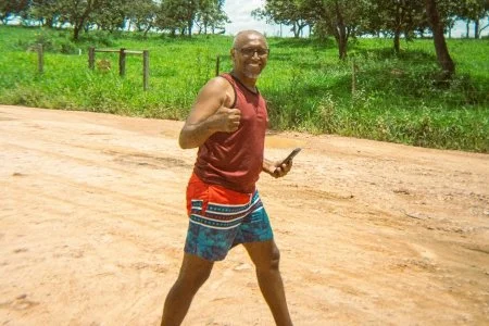 A smiling man in a red tank top and colorful shorts jogging on a dirt path in a green park