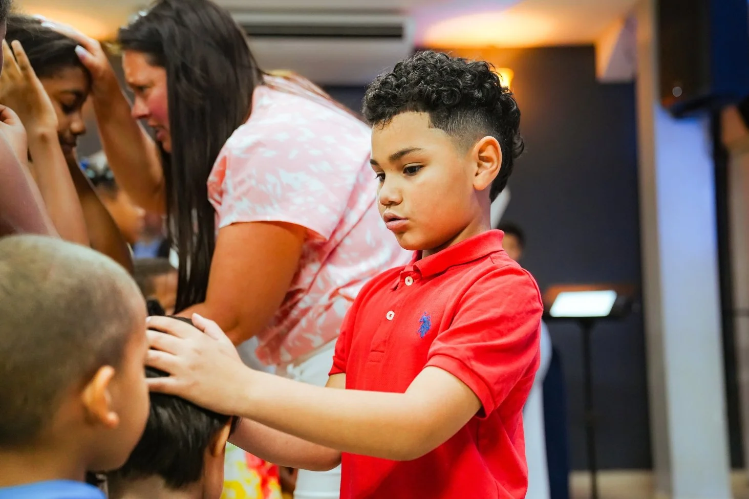 A young boy with curly hair and wearing a red polo shirt is praying, placing his hands on the head of a girl with short hair in a group prayer setting, with other children and an adult woman participating.