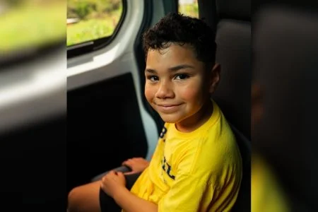 Young boy wearing a yellow shirt sitting on a bus with a window behind him.