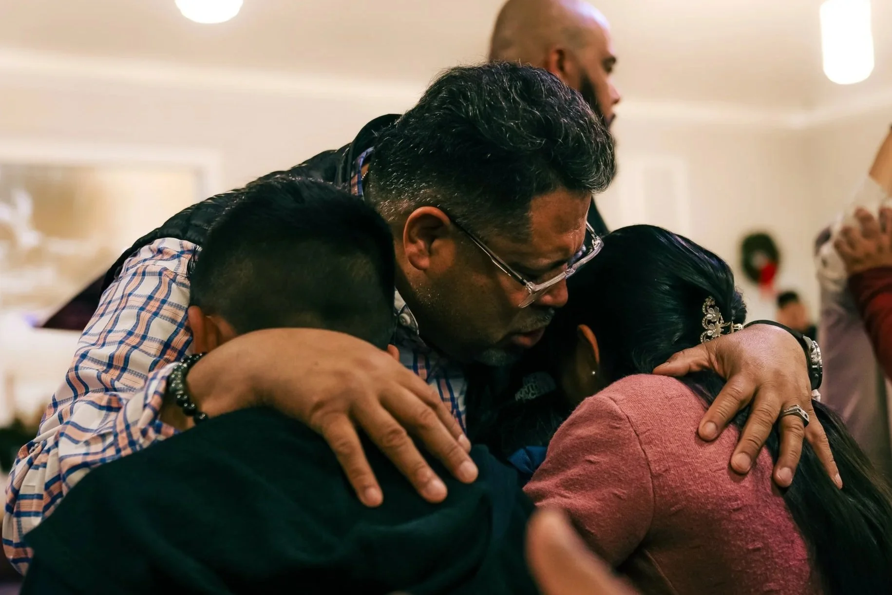 People hugging in a group at an indoor gathering, with one man wearing glasses and a checkered shirt, all showing expressions of comfort and compassion.