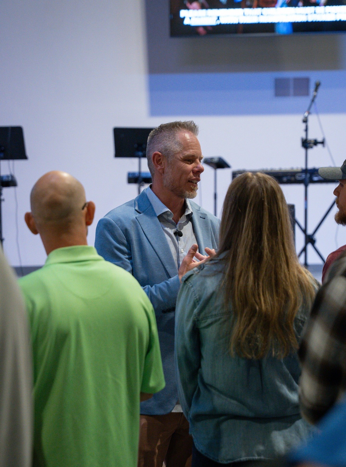 A middle-aged man with gray hair and beard, wearing a light blue blazer and white shirt, speaking to a group of four people in a room with musical equipment and a wall-mounted screen in the background.
