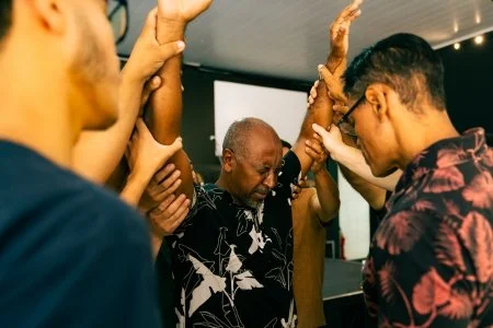 Group of people holding hands up together in a circle, with a man in the center participating in the activity.