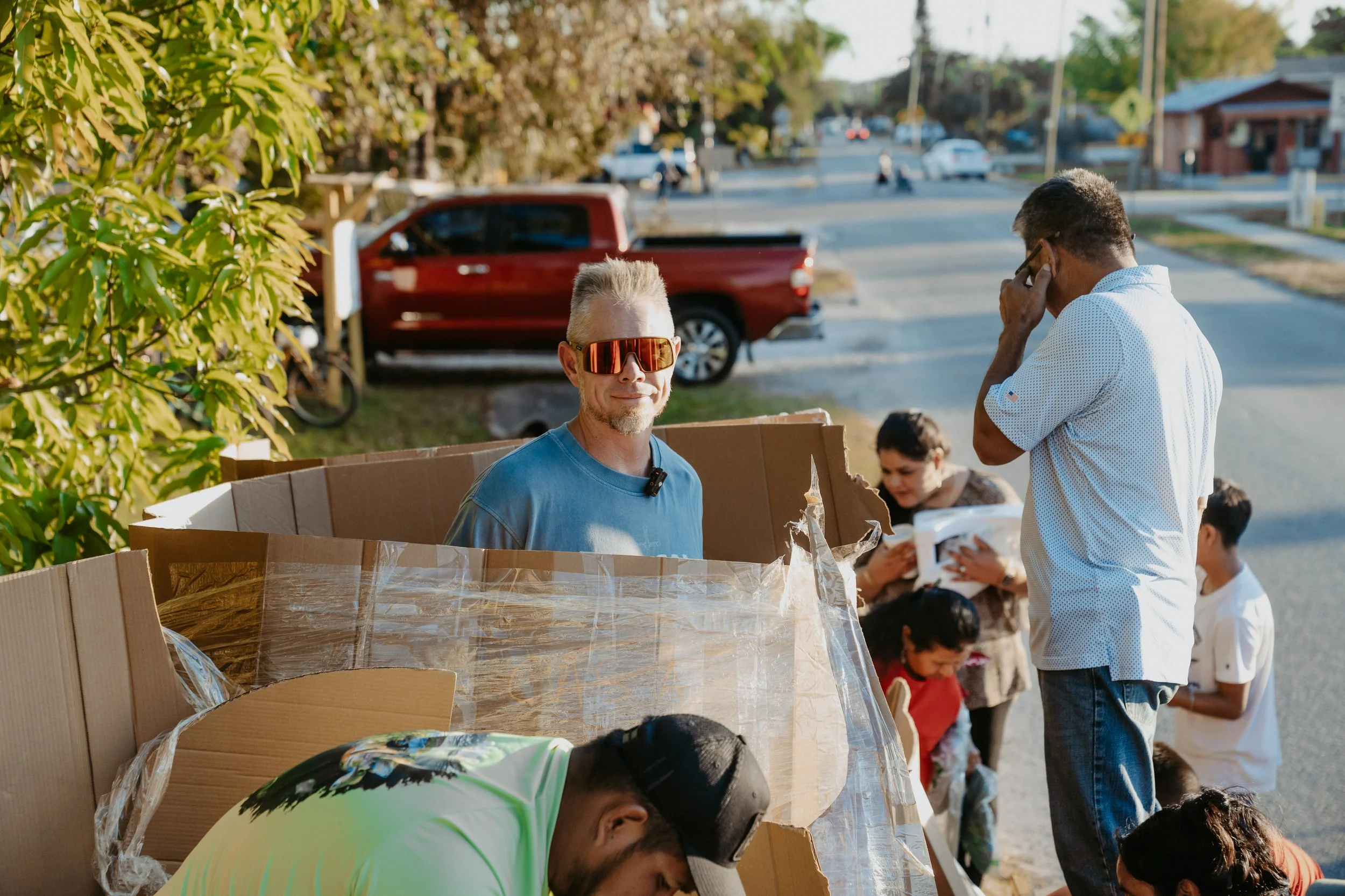People, including children and adults, gather outdoors near a row of cardboard boxes wrapped in plastic, with some children in line and a man resting his head on a box, a man in sunglasses smiling, in a neighborhood with trees and vehicles in the background.