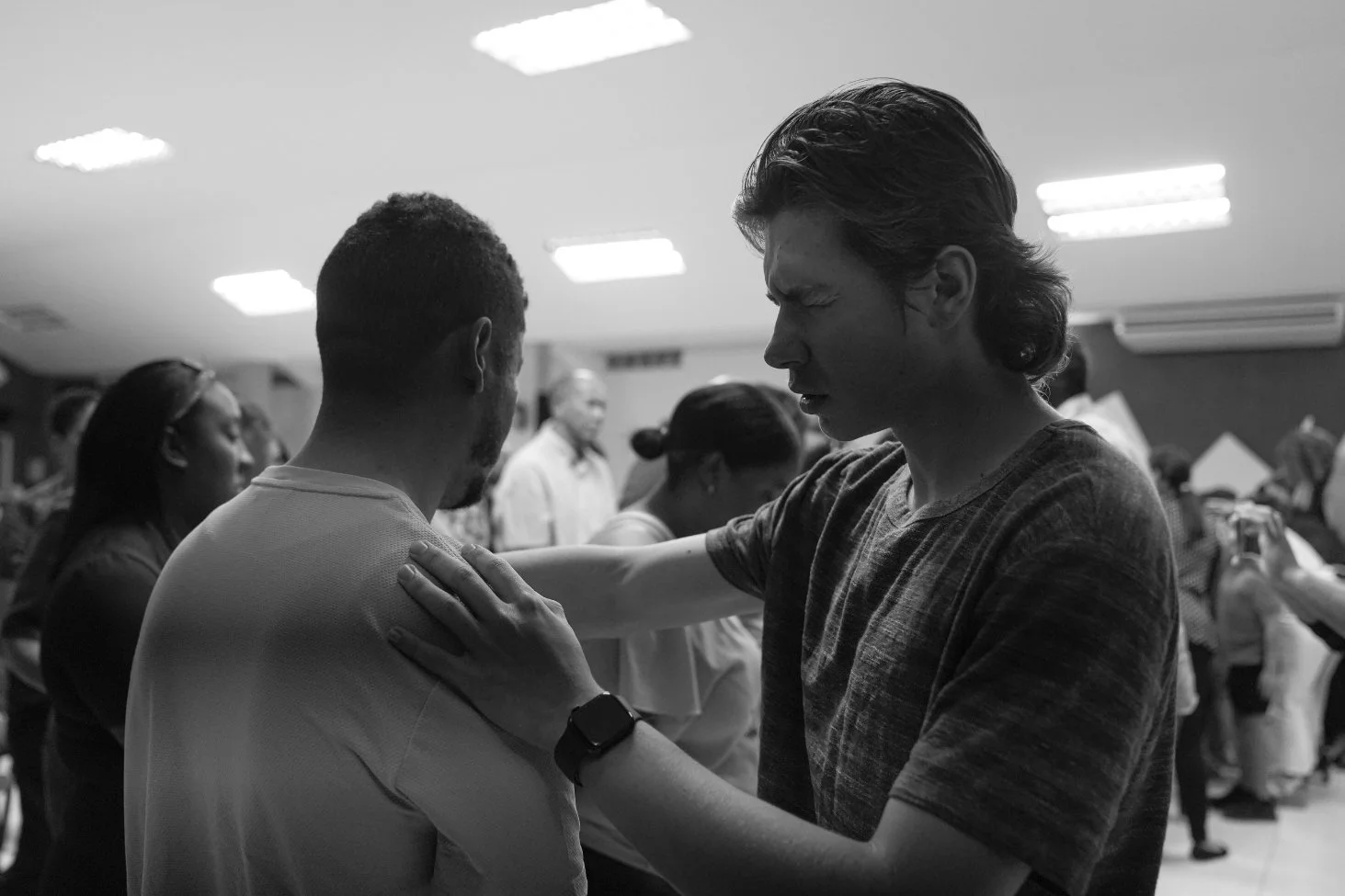 A man praying with a woman in a church, standing behind him with other people in the background.