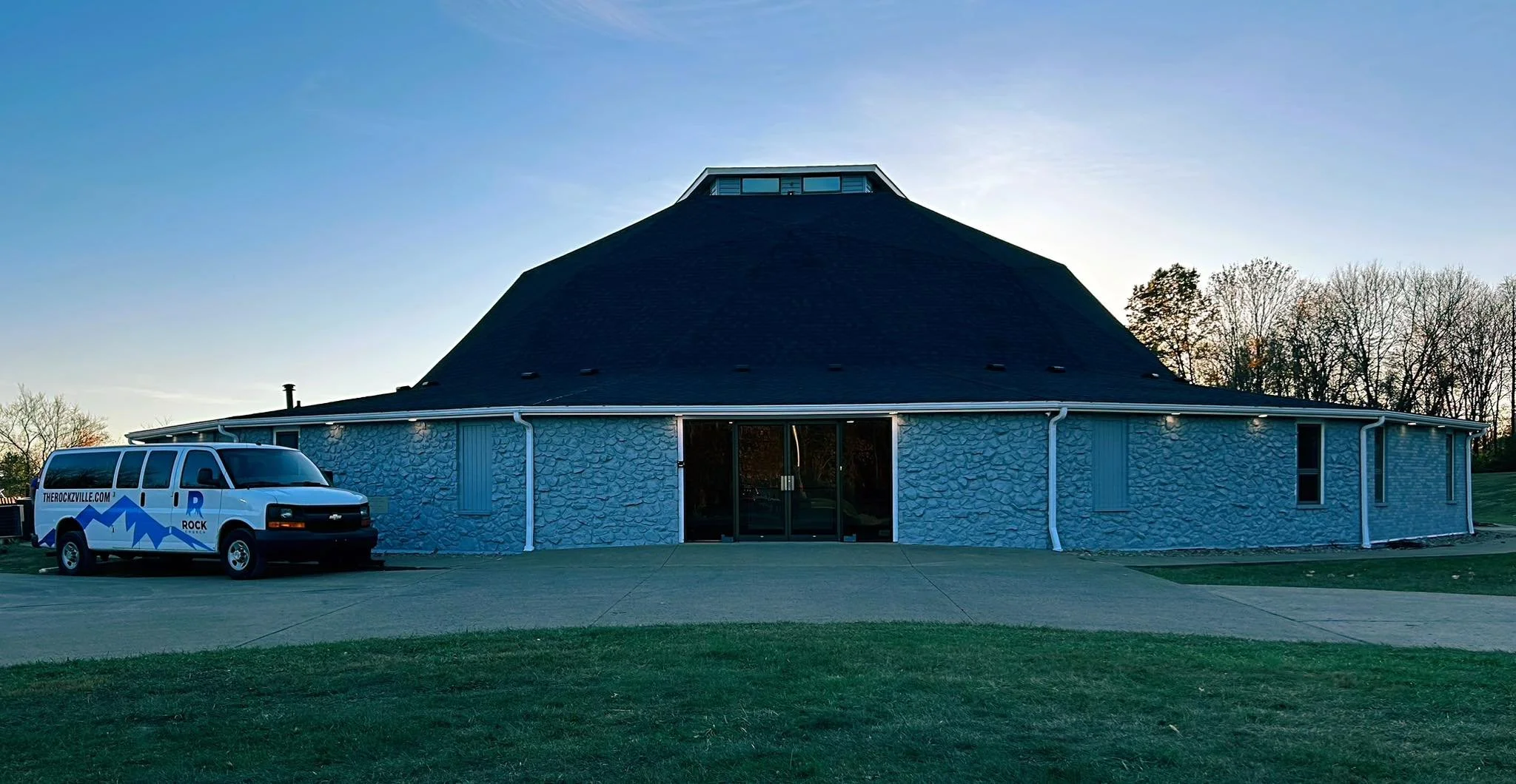 A round, castle-like building with a dark conical roof and light-colored stone walls, with a white van parked in front, there are trees in the background and the sky is clear.