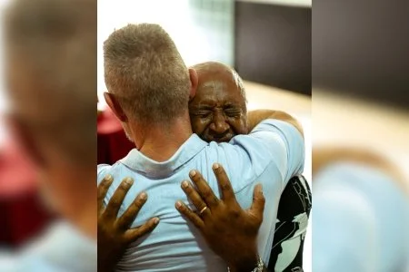 Two men hugging, one with gray hair and the other with short hair, inside a room.