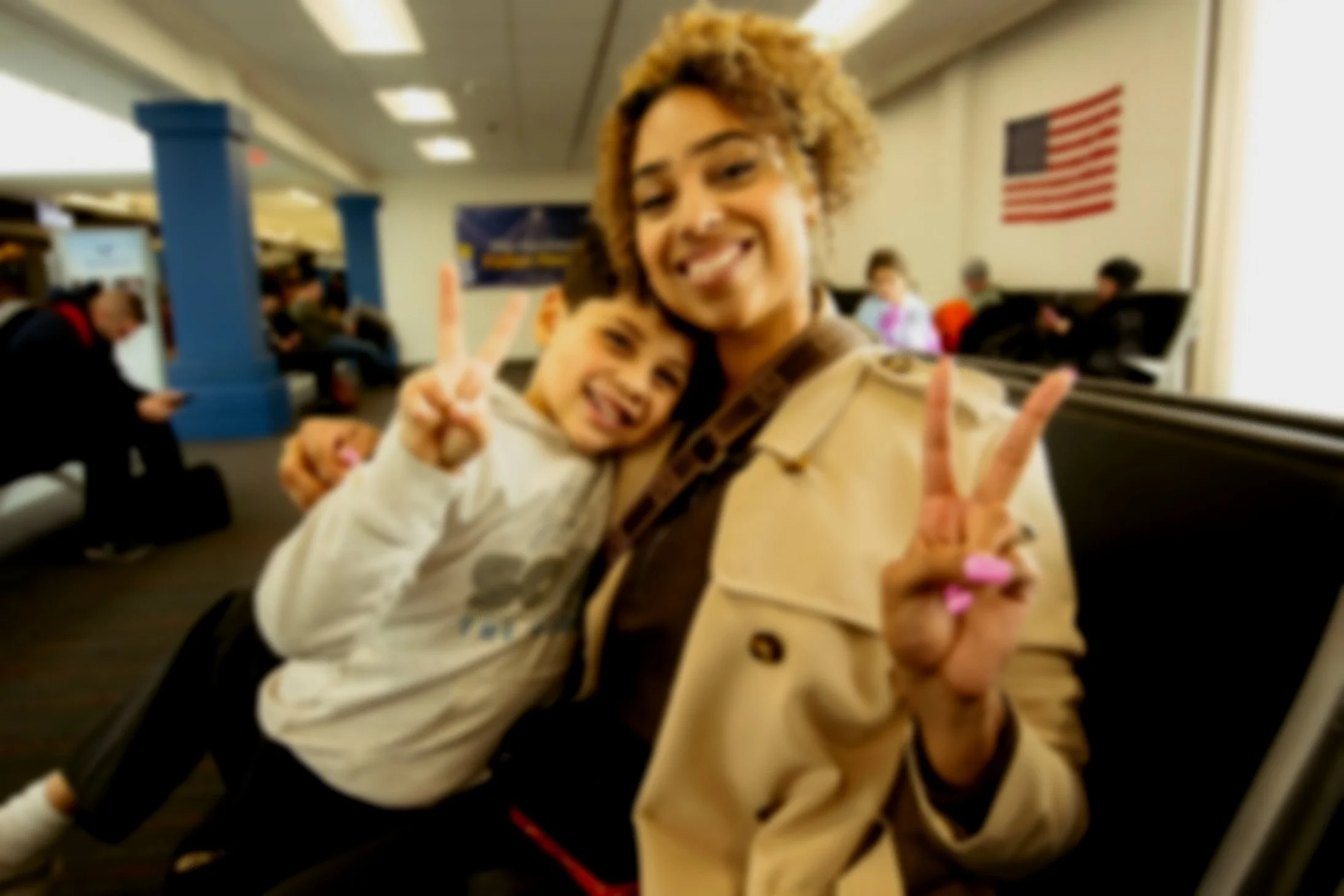A smiling woman and a girl posing for a selfie at an airport, with an American flag on the wall behind them.