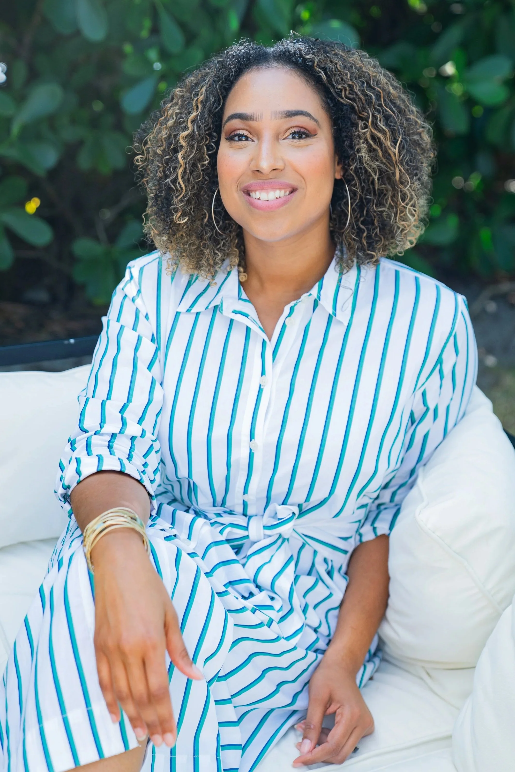 A young woman with curly hair and hoop earrings smiling while sitting on a white sofa outdoors in front of green foliage. She is wearing a white and blue striped shirt.