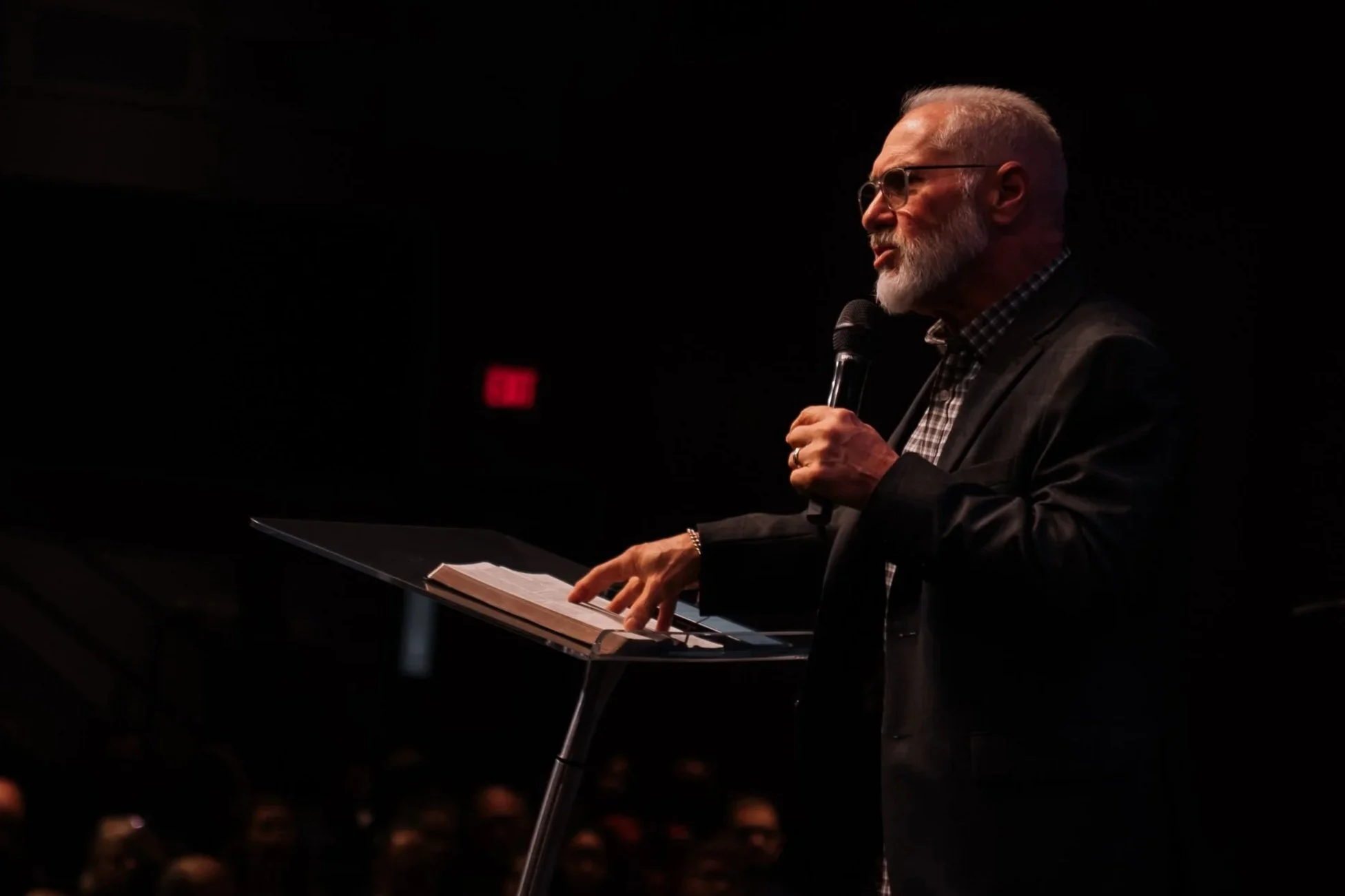 An older man with a beard and glasses giving a speech in a dimly lit auditorium. He is holding a microphone in one hand and resting the other on a music stand with papers. The man is dressed in a checkered shirt and a dark blazer.