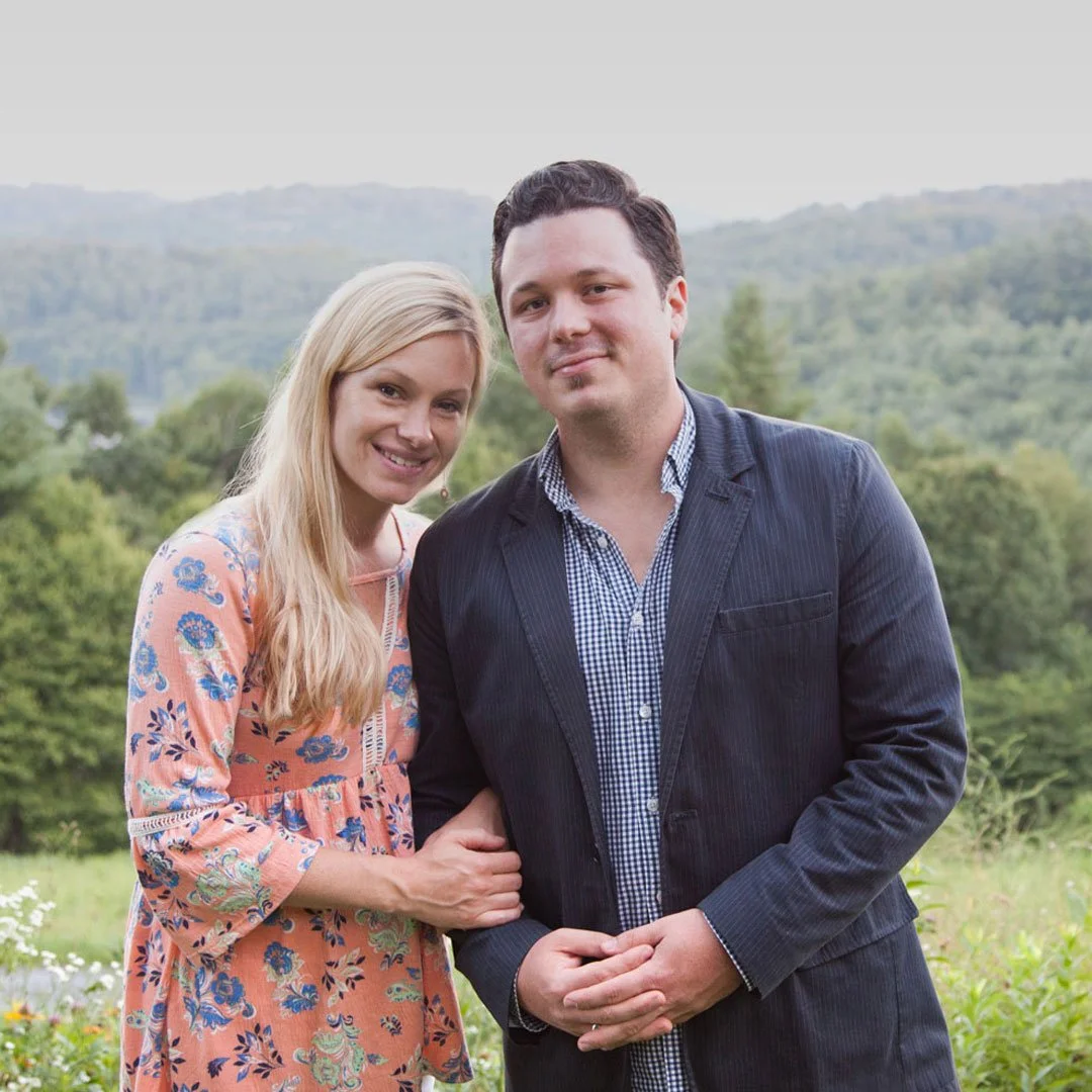 A couple standing closely together outdoors in a scenic green landscape with rolling hills in the background.