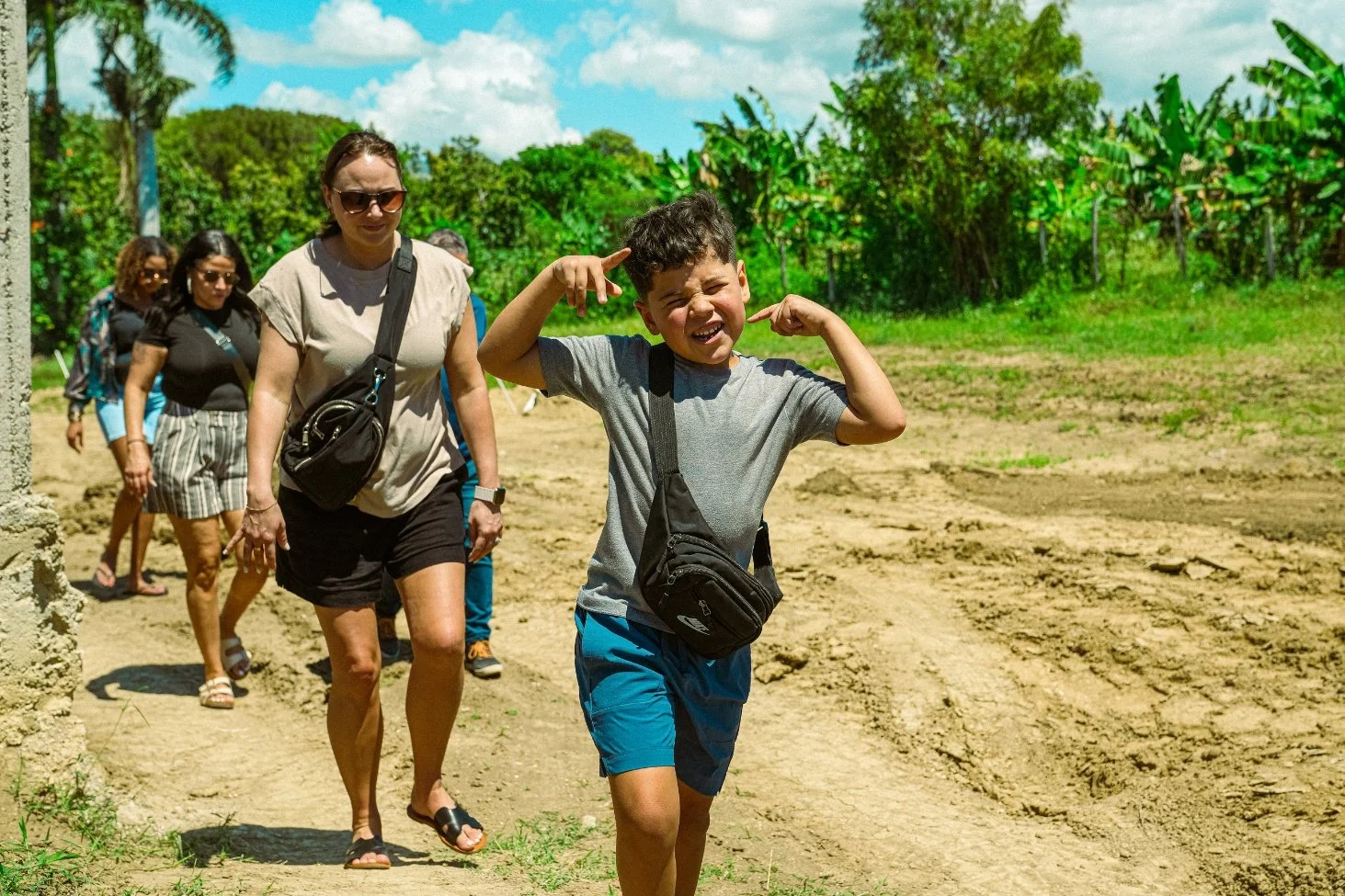 A group of people, including a boy in the front smiling and pointing at his head, walking on a dirt path in a lush green outdoor setting with trees and blue sky.