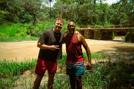 Two men standing outdoors near a small river, with lush green trees in the background. One is pointing at the other, and they are smiling.