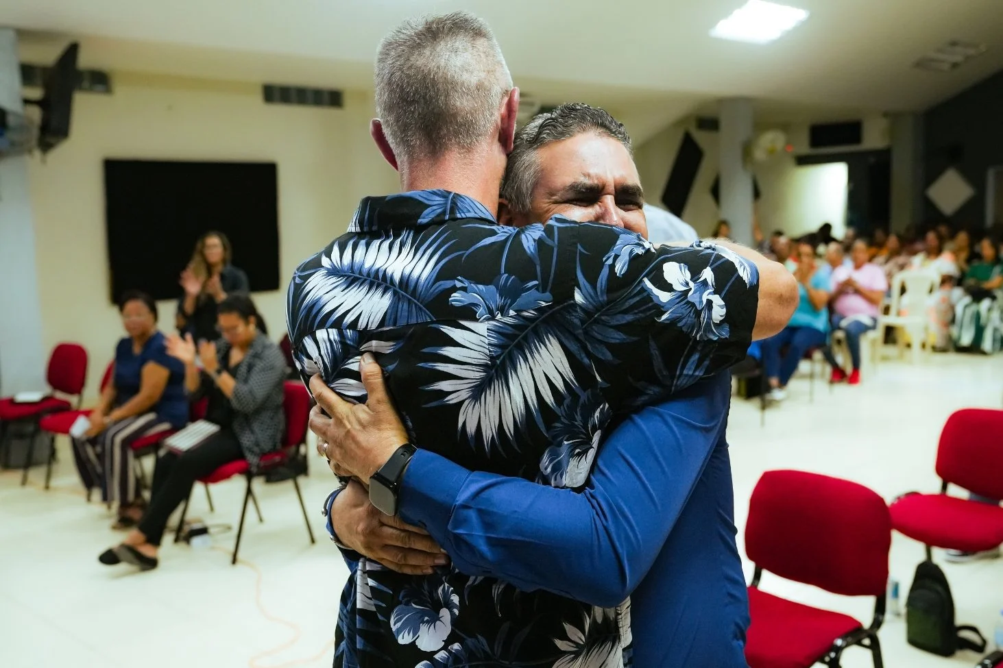 Two men hugging each other in an emotional embrace at a gathering, with people seated and clapping in the background.