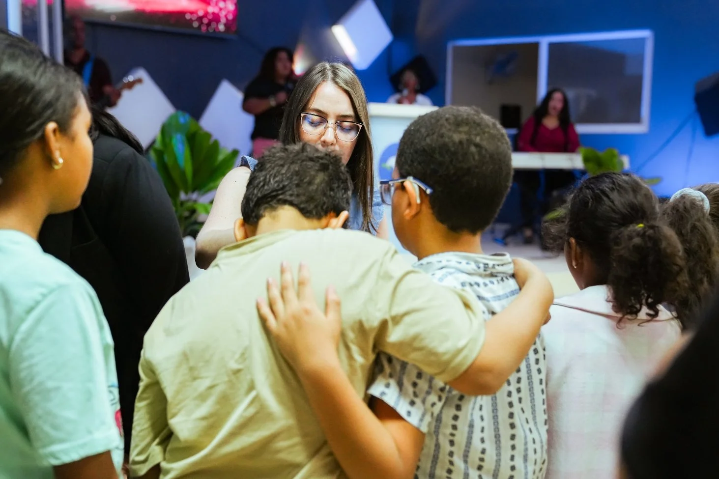 A group of children gathered closely around a woman in a room with blue walls, a large screen, and musical equipment, possibly during a group activity or discussion.