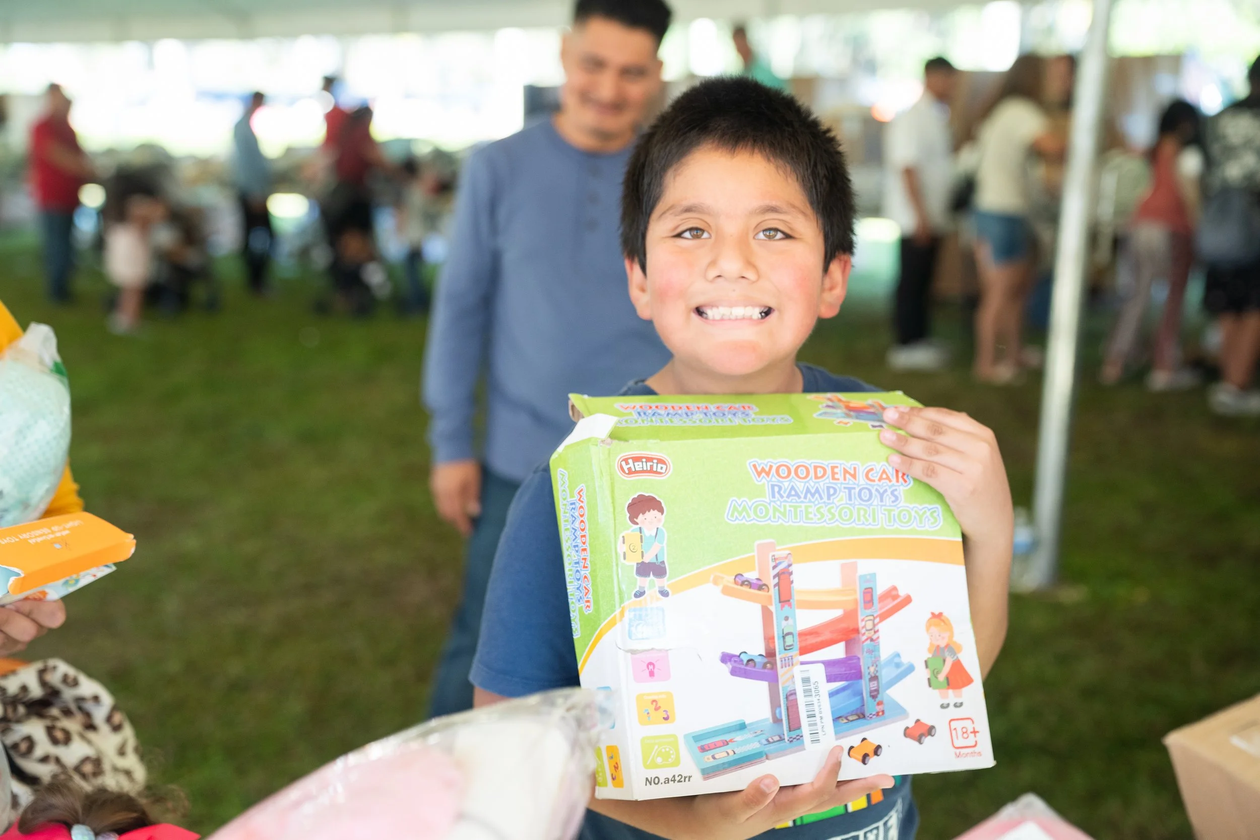 A young boy holding a colorful toy building set box smiling at the camera at an outdoor event, with a man standing behind him and a blurred background of people and activity in a park or covered pavilion.