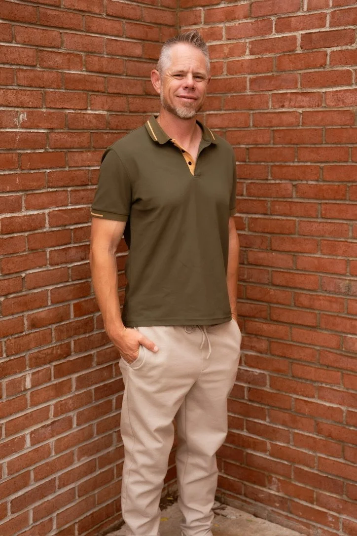 A middle-aged man with short gray hair, a goatee, wearing a green polo shirt with yellow collar and sleeve accents, beige jogger pants, standing against a red brick wall.