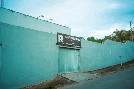 A blue concrete wall with a large garage door and a sign that reads 'IRESPROCHA' in front of a street, with a hillside and greenery in the background.