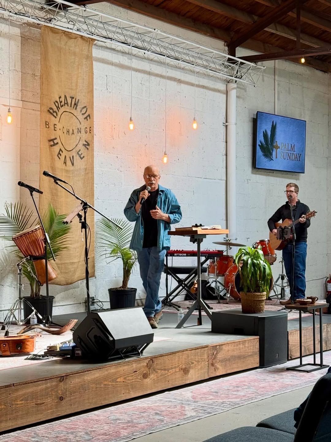 A man singing with a microphone on a stage, accompanied by a guitarist. Stage has musical instruments, plants, and a large banner with the words 'Breath of Heaven' and 'Be Change.' A screen reads 'Palm Sunday' with a palm leaf and cross symbol.