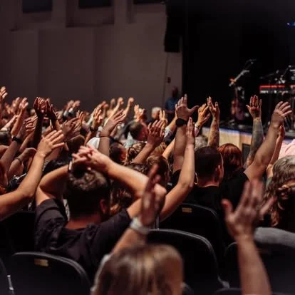 Audience raising hands during a live event or concert in a dark indoor auditorium.