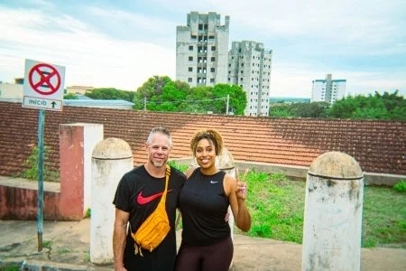A man and woman standing outdoors in front of a brick wall, with tall buildings in the background. The man is wearing a black shirt with a red swoosh and has a yellow bag across his shoulder. The woman is wearing a black athletic top and pants, smili