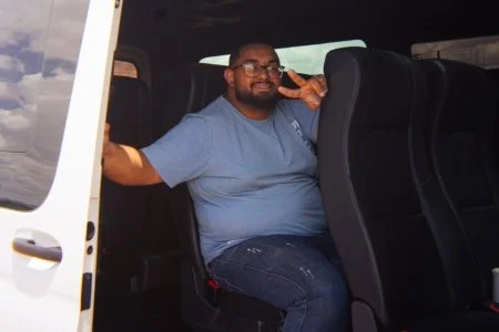 A man sitting inside a vehicle, smiling, and making a peace sign with his hand.