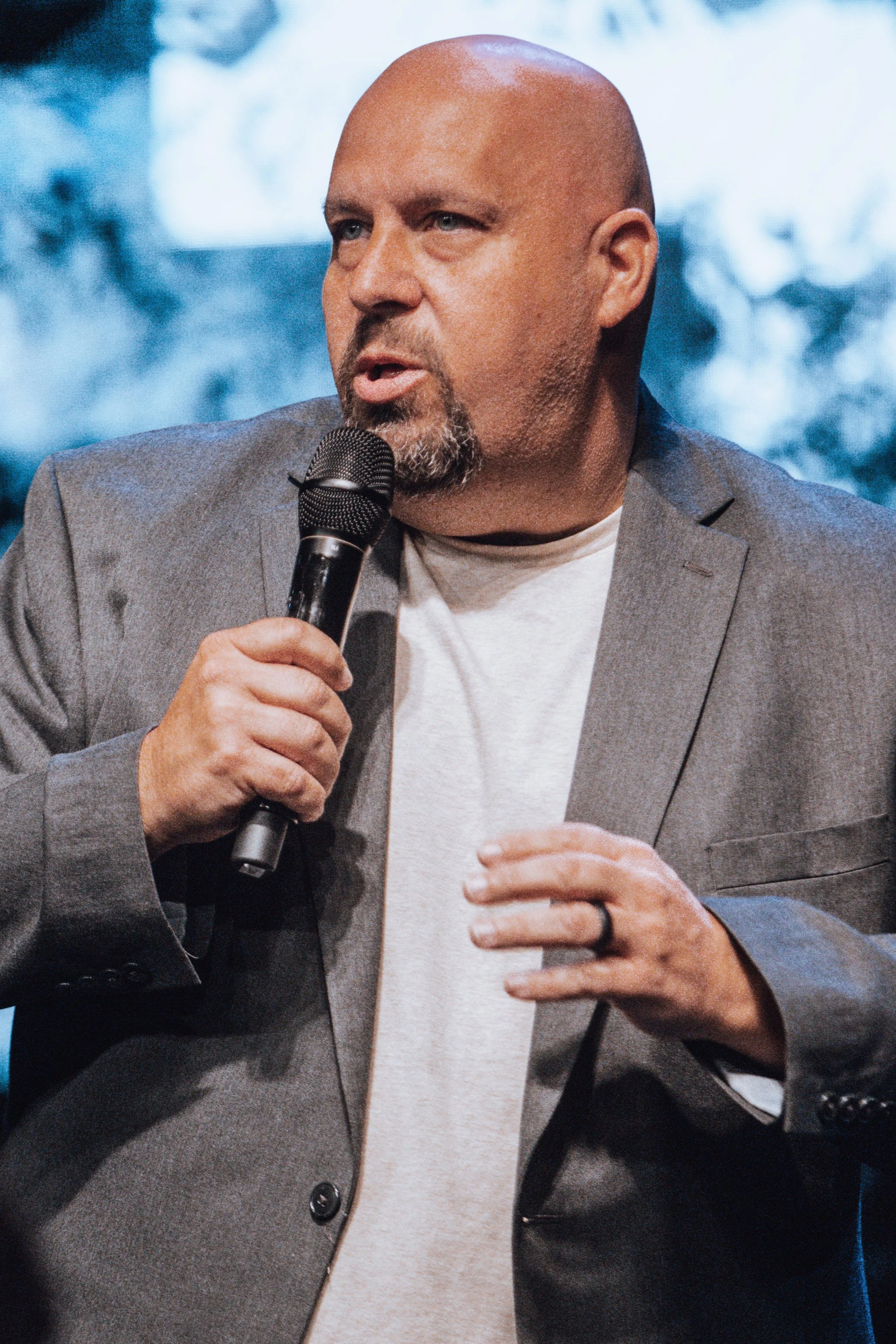 A man with a beard and a shaved head is holding a microphone and speaking, wearing a gray blazer over a white t-shirt, with a blurred background.