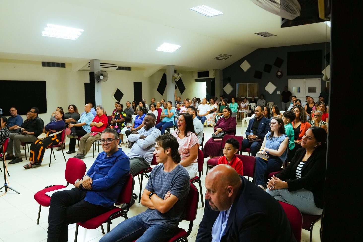 An audience seated in chairs attending a presentation or event in a large room, with some smiling and focusing on the speaker.