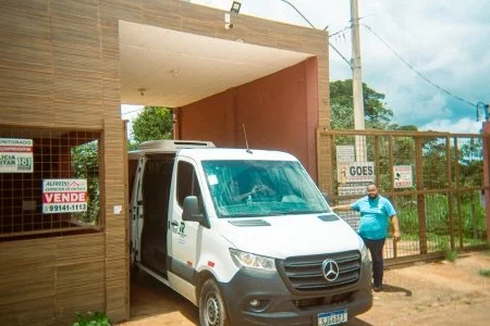 A white Mercedes-Benz van parked in front of a building with a man standing nearby. The building has a sign indicating it is for sale, and there is a gate and fence surrounding the area. Power lines are visible in the background under a partly cloudy