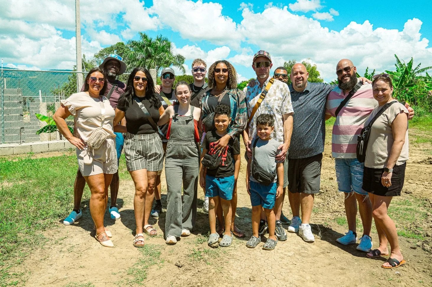 Group of 16 diverse people smiling outdoors on a sunny day, with greenery and a fence in the background.