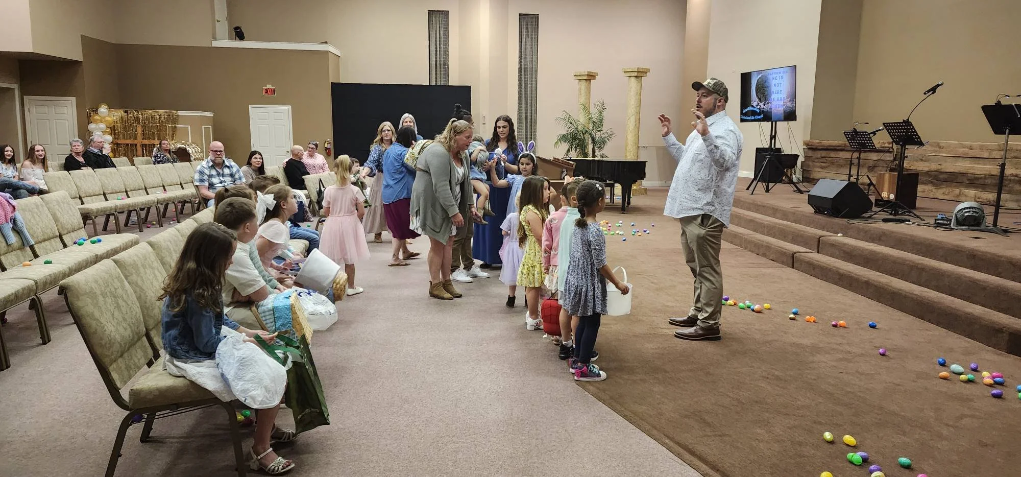 Children and adults participating in an Easter egg hunt inside a church or community hall. Children are lined up with baskets, and eggs are scattered on the floor.