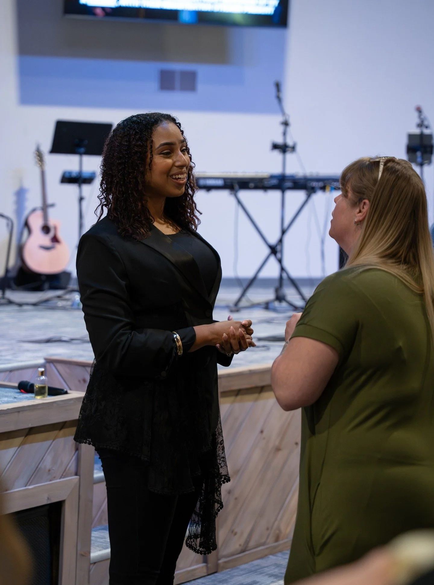 Two women are engaged in a conversation indoors, with musical instruments like a guitar and keyboard in the background, and a large screen on the wall.