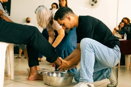 Two men performing a foot washing ritual with a basin of water, surrounded by a group of people in a room.