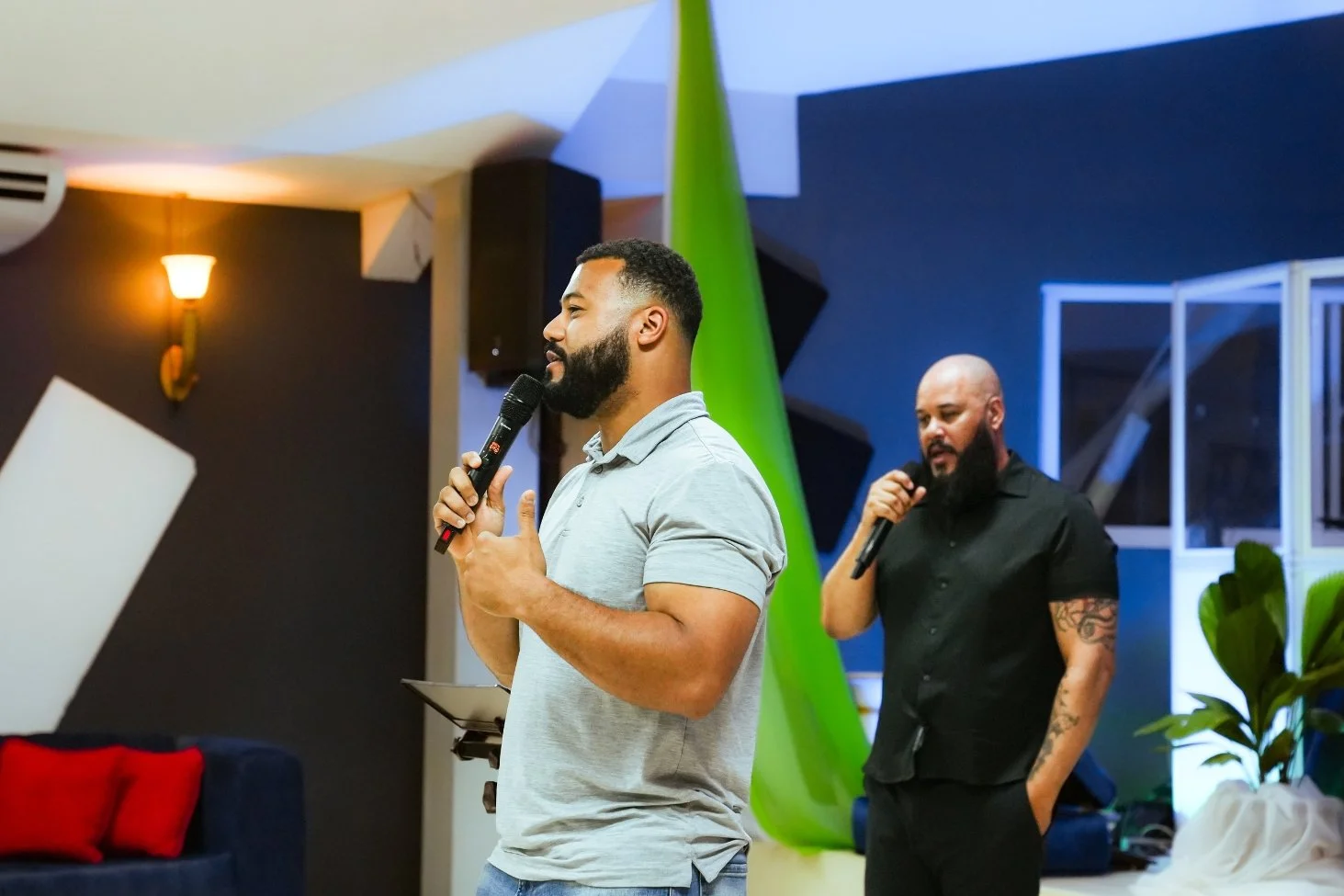 Two men speaking into microphones at an indoor event. One man in the foreground with a beard and wearing a gray polo shirt, the other in the background with a tattooed arm wearing a black shirt.
