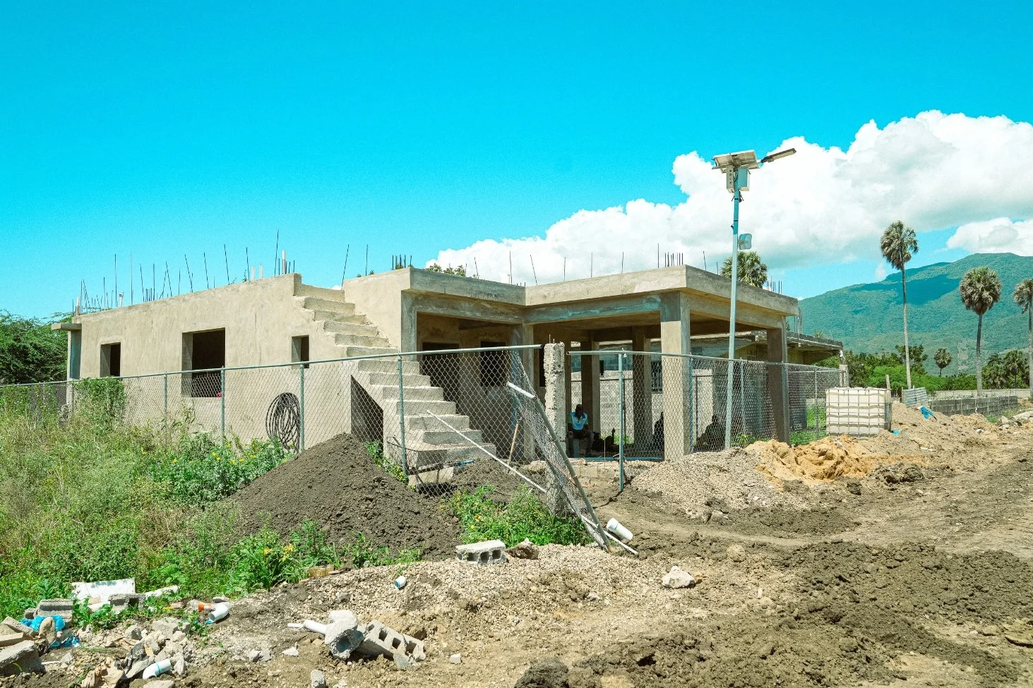 Under-construction building with concrete stairs, chain-link fence, construction dirt, and construction worker sitting outside. Mountain and palm trees in background, clear blue sky.