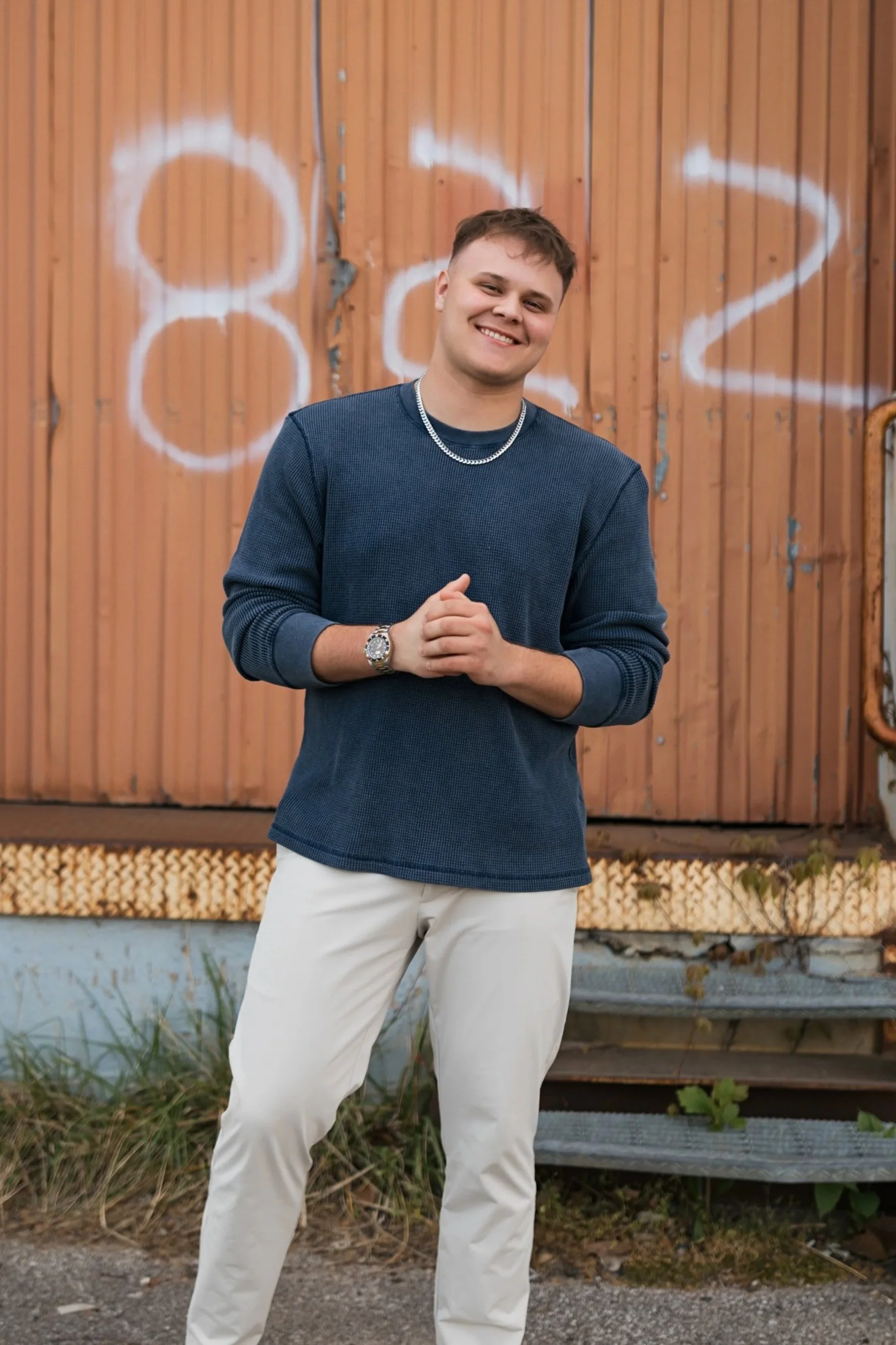 A young man standing outdoors in front of a wooden wall with white graffiti, smiling and wearing a navy blue sweater, white pants, a silver chain, and a wristwatch.