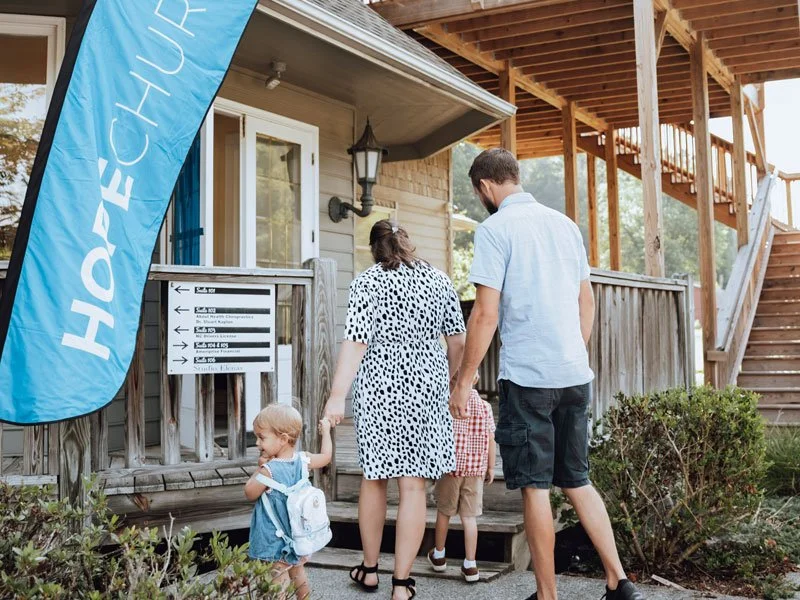 Family with small children walking up to a building with a wooden exterior, stairs, and a large blue flag that says 'HOPE CHURCH'.
