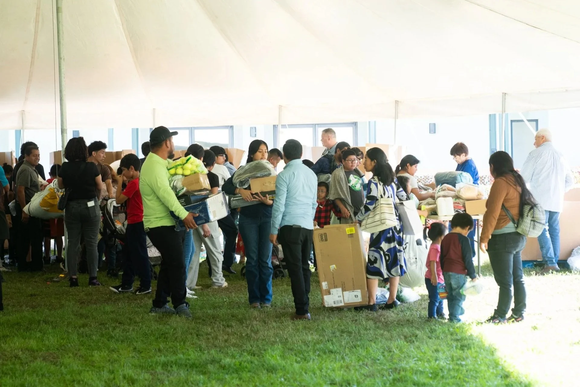 People in line at a distribution event under a large white canopy, with tables laden with supplies, including boxes and bags, on a grassy area.