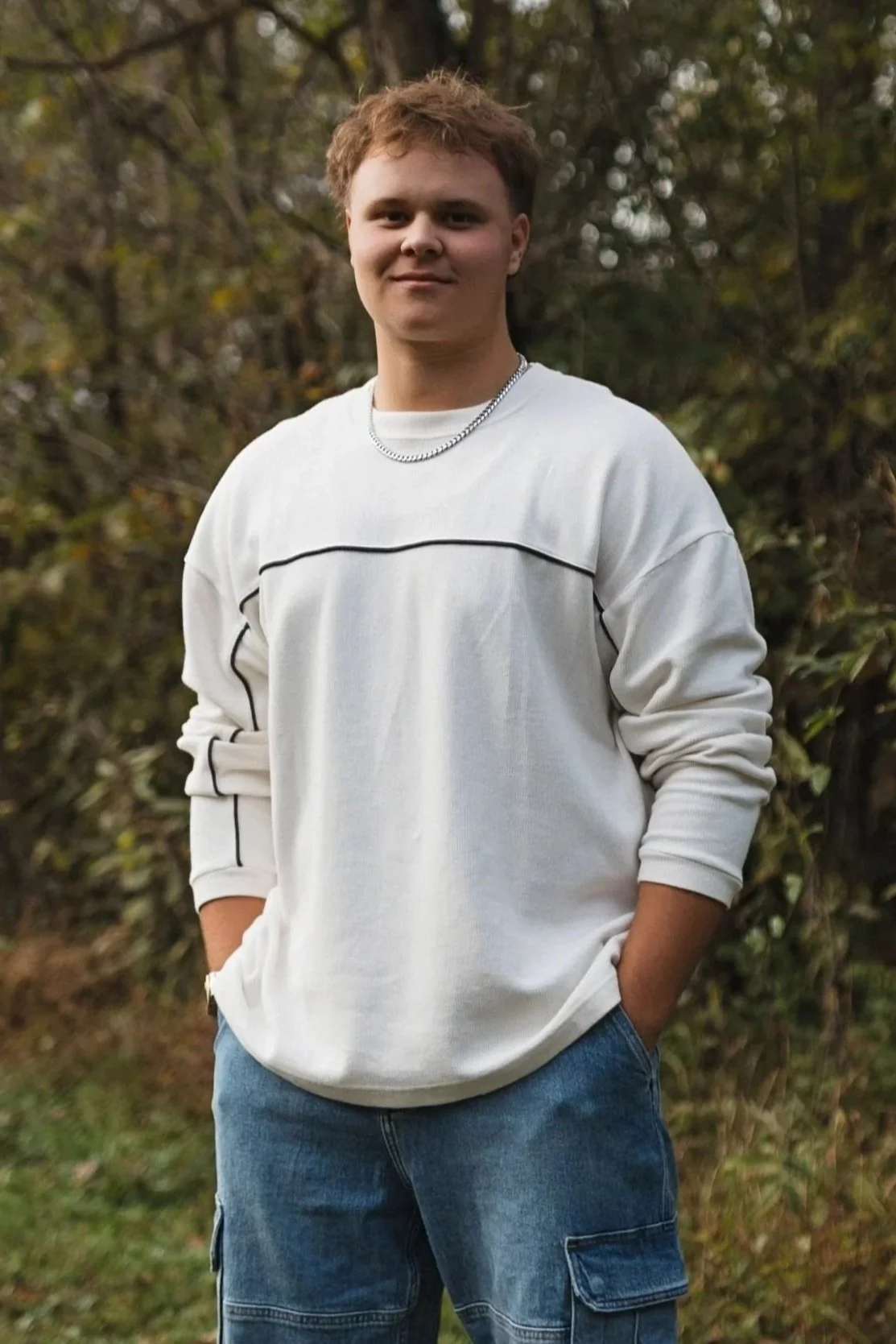 A young man standing outdoors in front of trees, wearing a white sweatshirt with black lines and blue jeans, hand in pocket, smiling