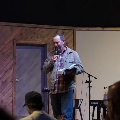 Man holding a microphone and reading from a book on stage in a wooden-paneled room.