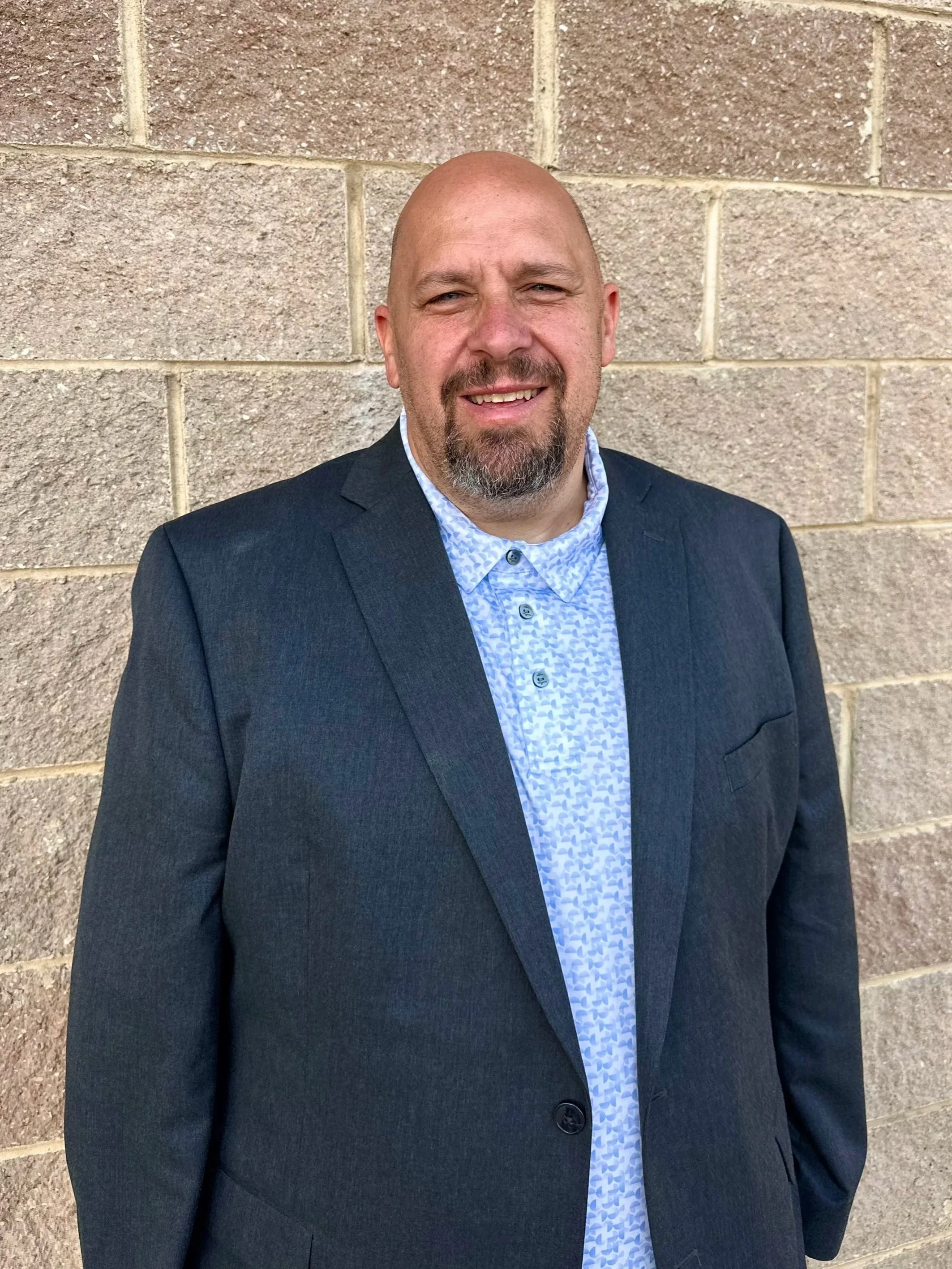 A man with a beard and balding head dressed in a dark blazer and light blue patterned shirt, standing against a brick wall, smiling at the camera.