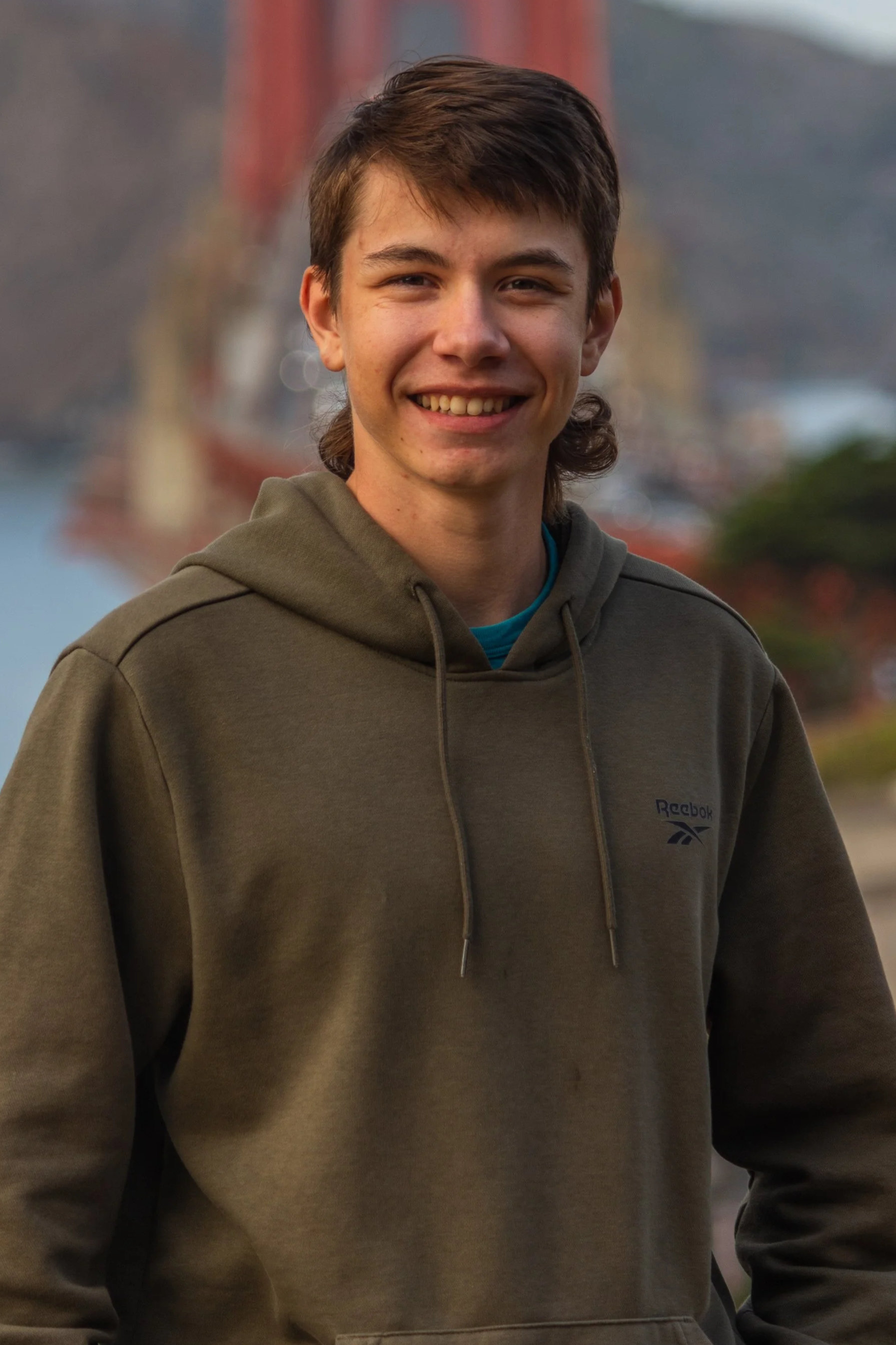 A young man with brown hair and fair skin, smiling, wearing a dark green Reebok hoodie, outdoors with a blurred bridge and water in the background.