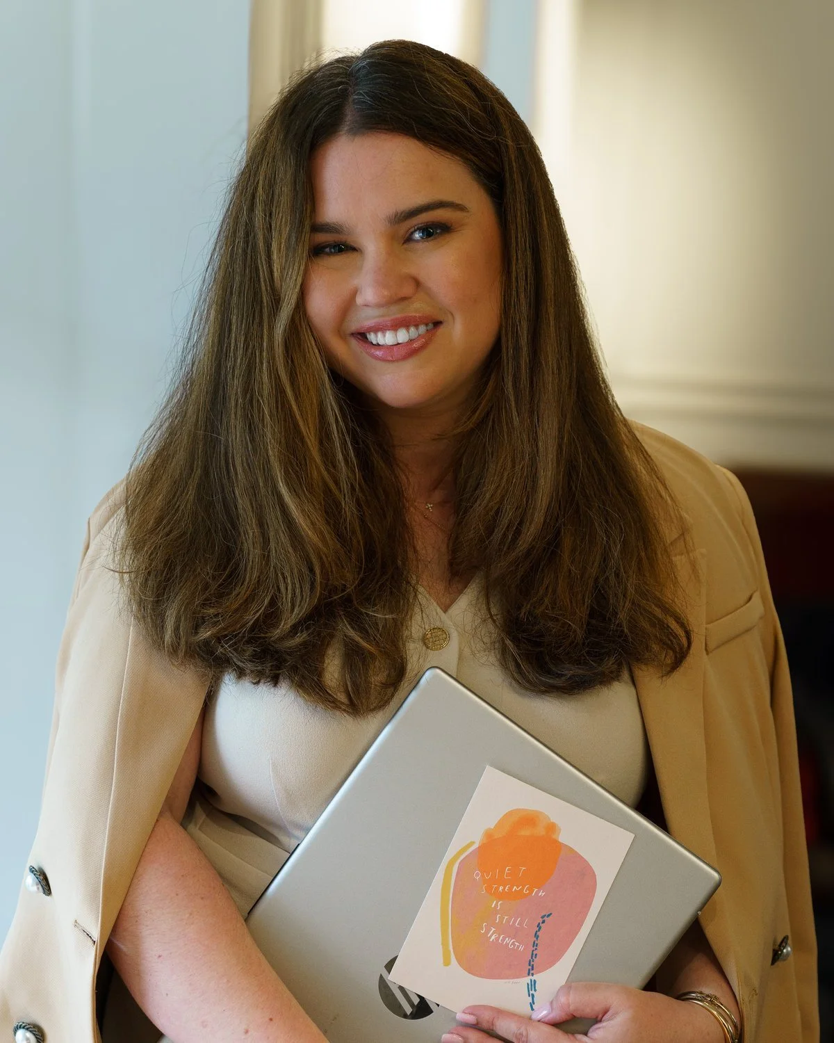 A woman with long brown hair and blue eyes smiling, holding a laptop with a colorful illustration and text about strength.