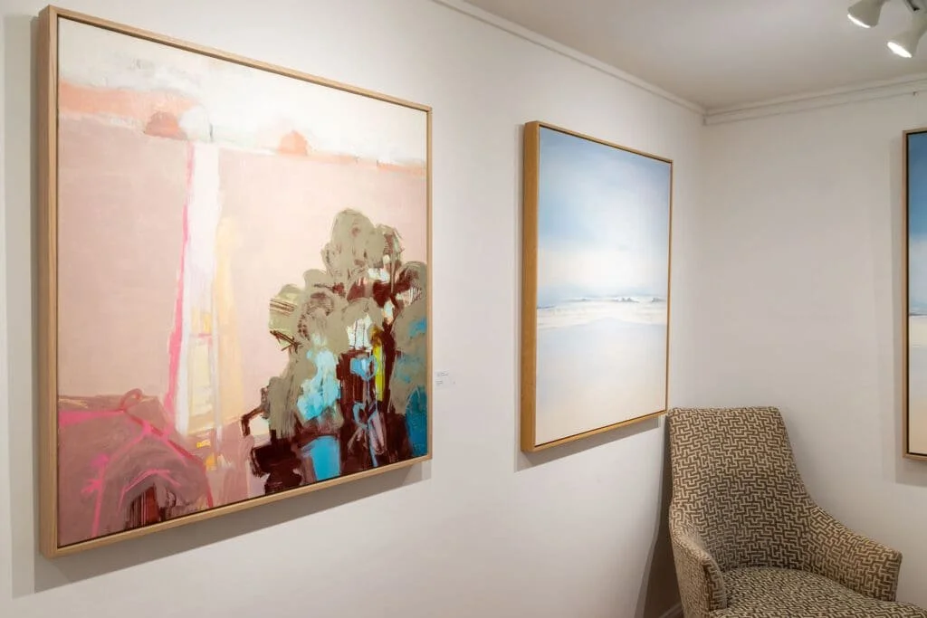 Three paintings hanging on a white wall in an art gallery, with a patterned armchair beside the paintings.