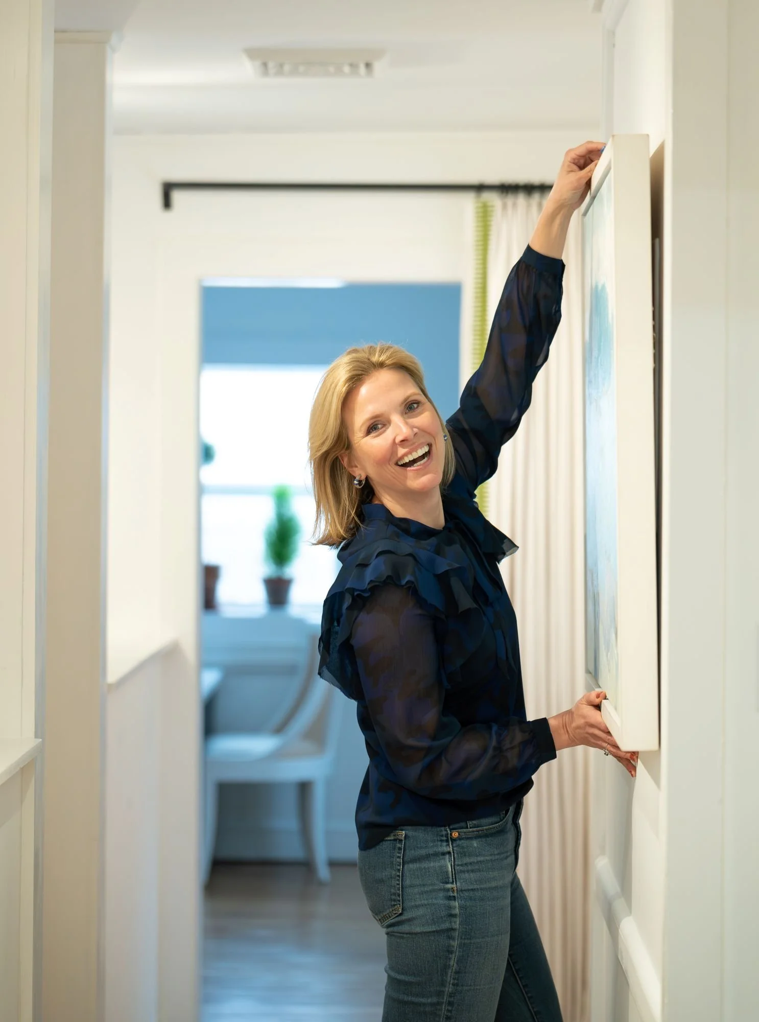A woman with blonde hair wearing a dark blue blouse and blue jeans, smiling, reaching for a thermostat or wall device in a hallway. The hallway has white walls, and there are curtains and a window in the background.