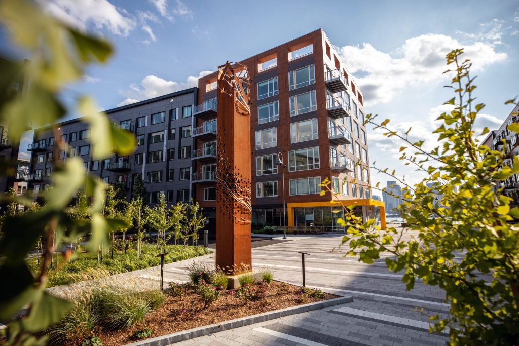 Modern apartment building with glass windows and balconies, surrounded by landscaped greenery and a paved walkway, under a partly cloudy sky.
