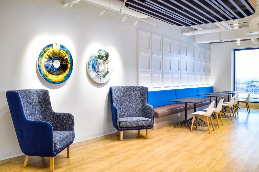 Modern interior with blue armchairs, a long blue bench with brown cushion, white chairs, tables, wall art, and large window showing cityscape.