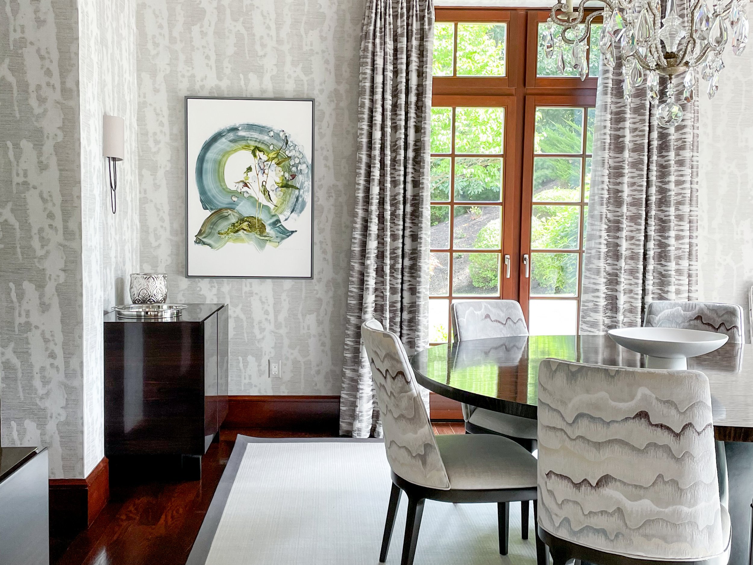Dining room with a round black table, upholstered chairs, large windows with curtains, a chandelier, a painting on the wall, and a side table with a glass decorative object.