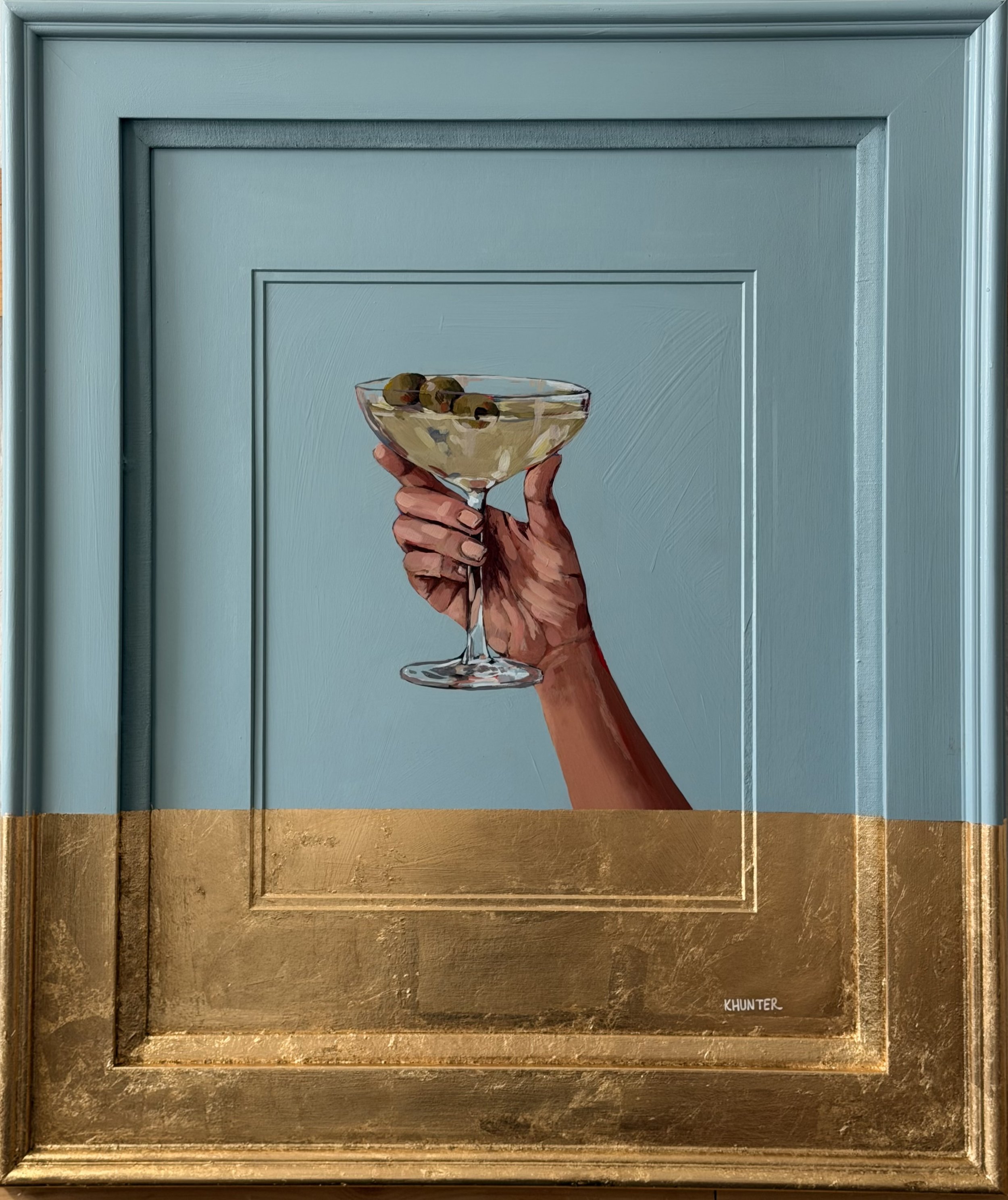 Painting of a hand holding a Martini glass with olives, signed custom signature 'KHUNTER' on the bottom right, mounted on a teal door with a gold frame at the bottom.