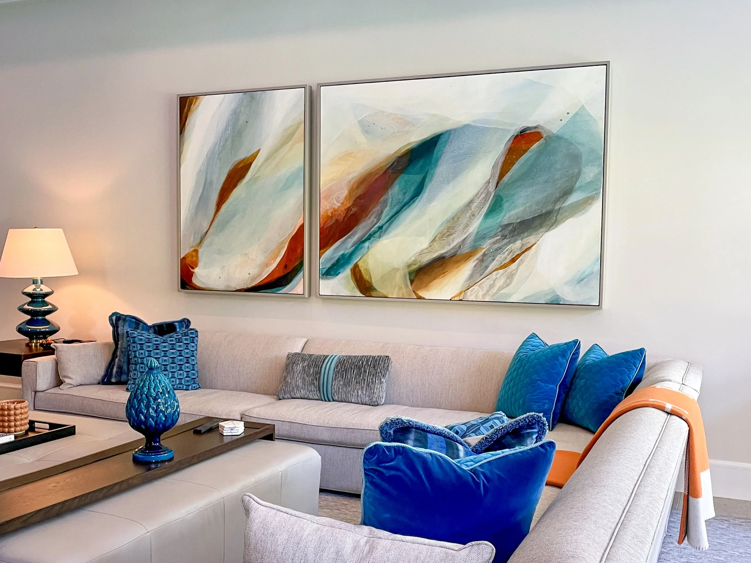 Living room with a beige sofa decorated with blue pillows, a wooden coffee table holding blue vases and other accessories, a lamp on a side table, and a large abstract painting hanging on the wall.