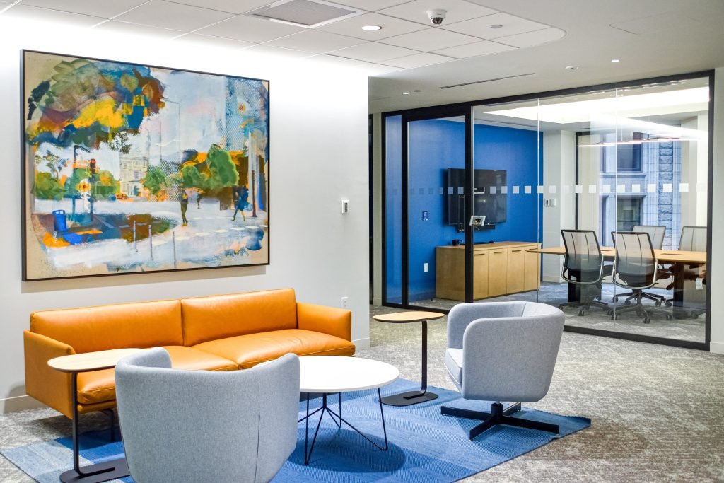 Modern office lobby with an orange sofa, two light gray armchairs, a small table, and an abstract painting on the wall, with a conference room visible through glass doors in the background.